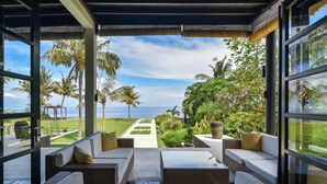 A covered outdoor seating area with wicker furniture and cushions, overlooking a well-maintained garden and the ocean. - Luxury Beach Front Villa with large garden, private pool, staff and free Wi-fi (Singaraja)