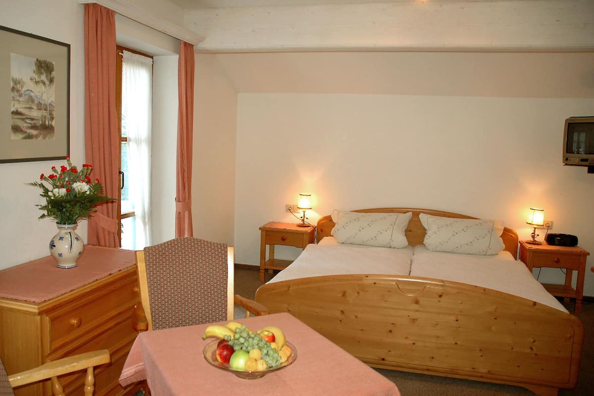 A bedroom with a wooden bed, two bedside lamps, a nightstand with a radio, a chair, a table with fruit, a vase with flowers, and a painting on the wall.