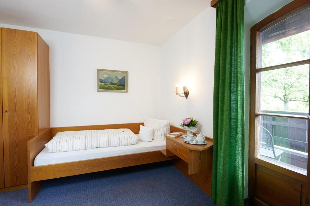 A single bed with a wooden frame, a nightstand with a vase of flowers, a wall-mounted lamp, and a window with green curtains.