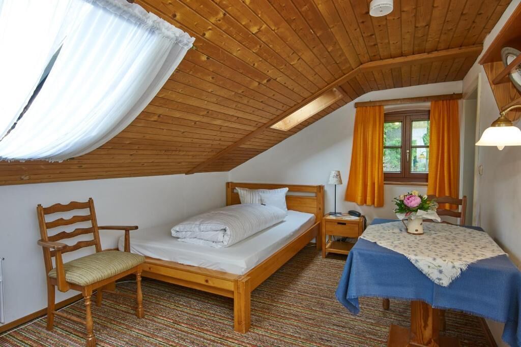 A cozy bedroom with a wooden ceiling, a single bed, a chair, a table with a tablecloth, and a window with curtains.