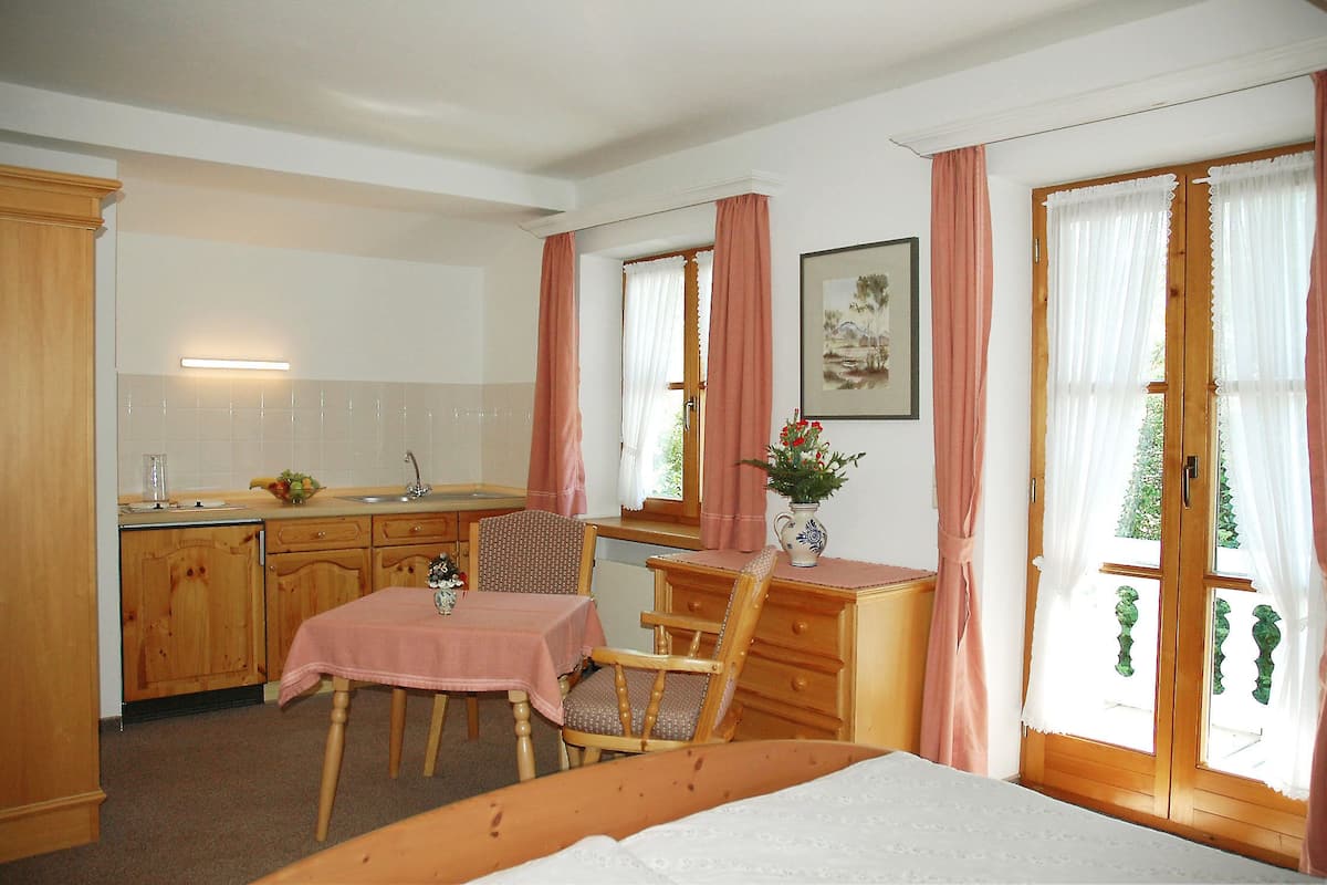 A compact room with a bed, a dining table with chairs, a kitchen area with wooden cabinets, and a window with curtains.