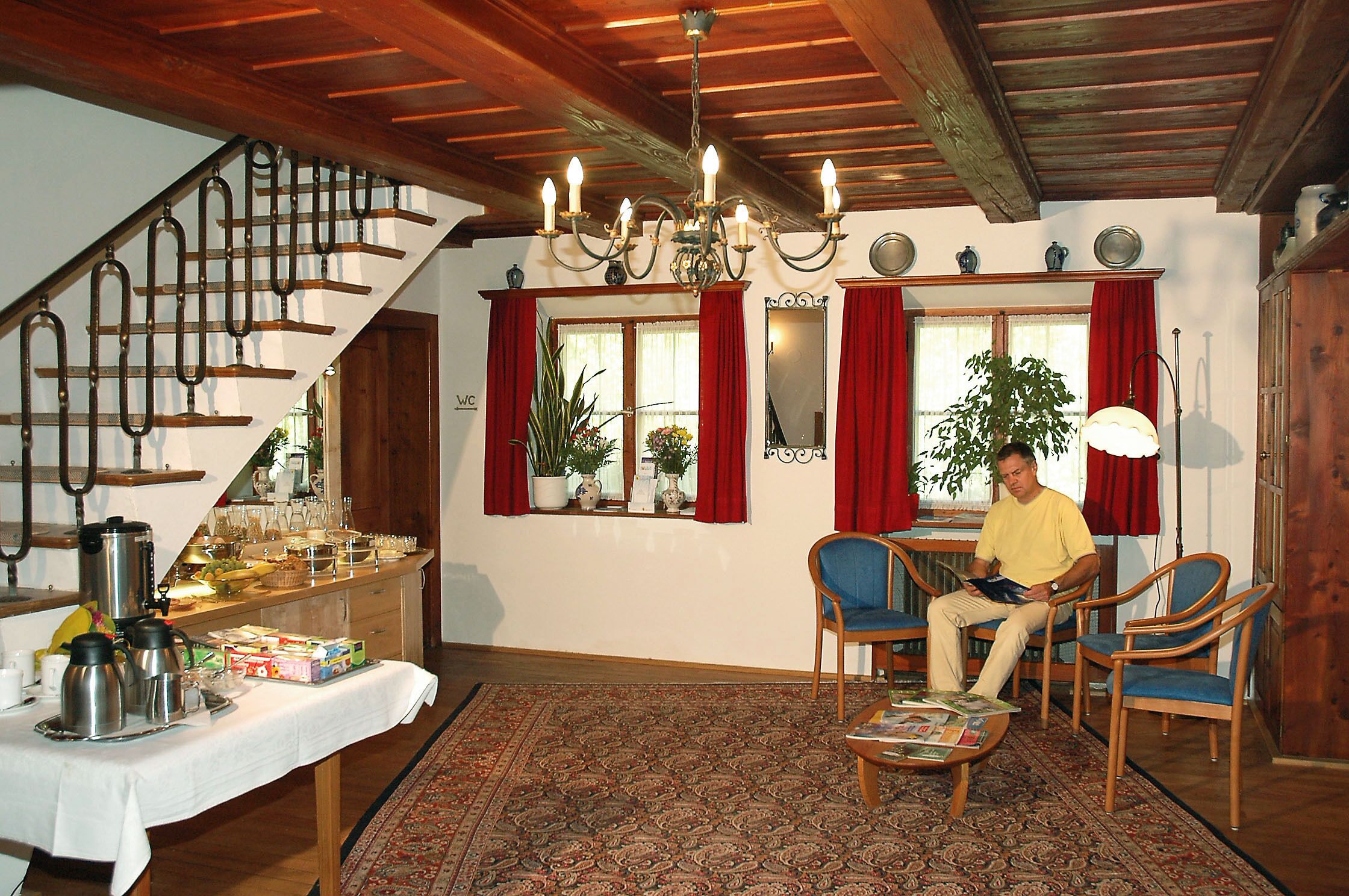 Uma sala de estar com escada, teto de madeira, um candelabro, um espaço de jantar com mesa posta para refeição e uma janela grande com cortinas vermelhas.