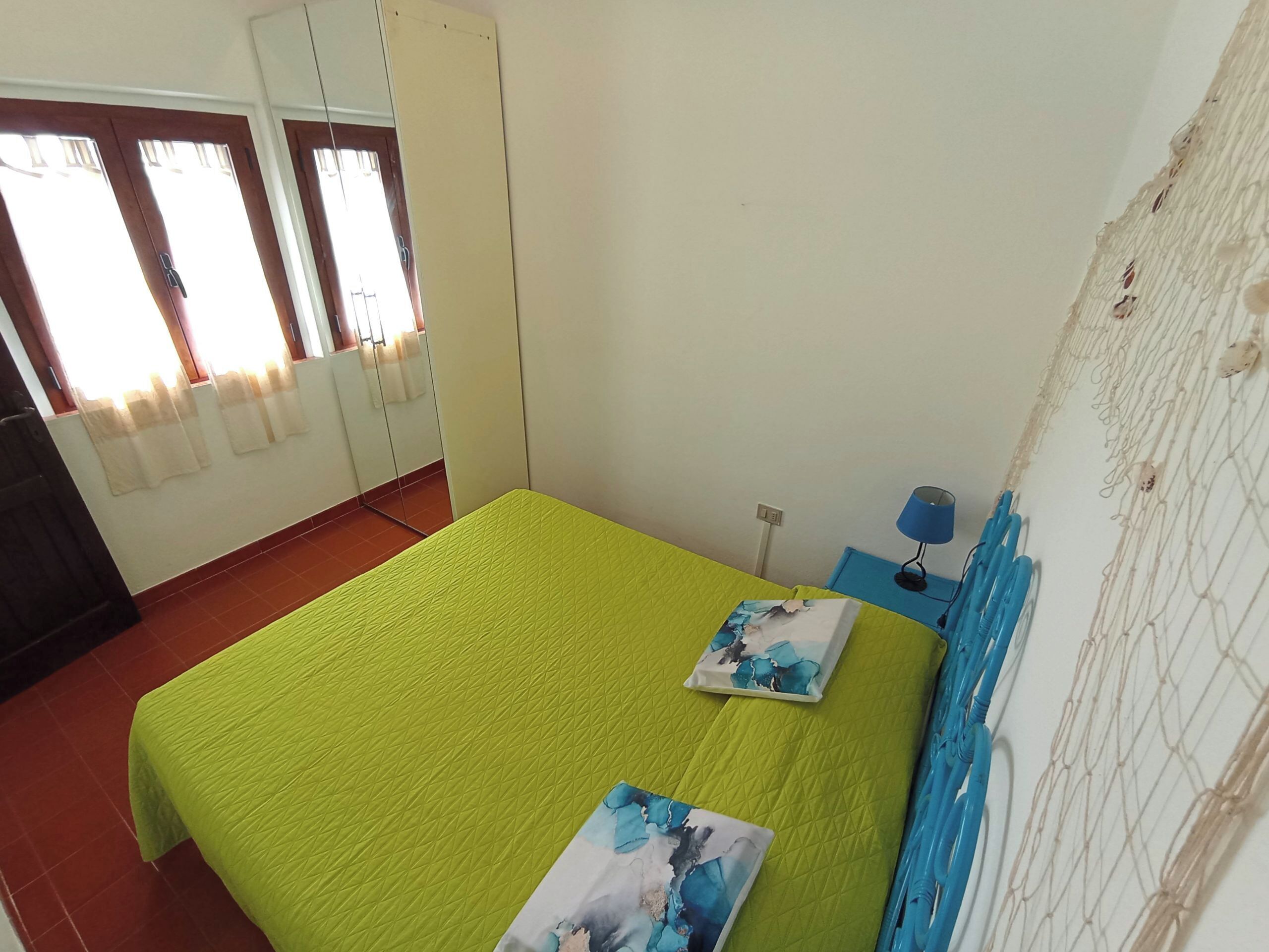 Una habitación con colcha verde, dos almohadas, una mesita de noche azul con lámpara y una ventana con cortinas.