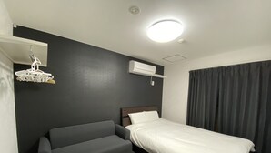 Ein kompaktes Hotelzimmer mit Bett, einer Schlafcouch, einem kleinen Tisch, einer Deckenleuchte, einer Klimaanlage und einer Pendelleuchte.