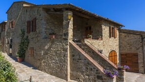 A stone building with a staircase leading to a wooden door, surrounded by potted plants and a clear blue sky.