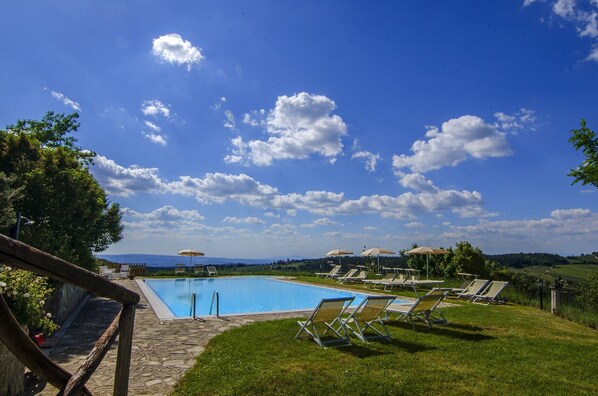 A swimming pool with lounge chairs and umbrellas, surrounded by greenery and a clear sky.
