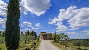 A rural road leading to a stone building with a tiled roof, surrounded by trees and a clear sky. - La Grotta Della Rocca (Castellina in Chianti)