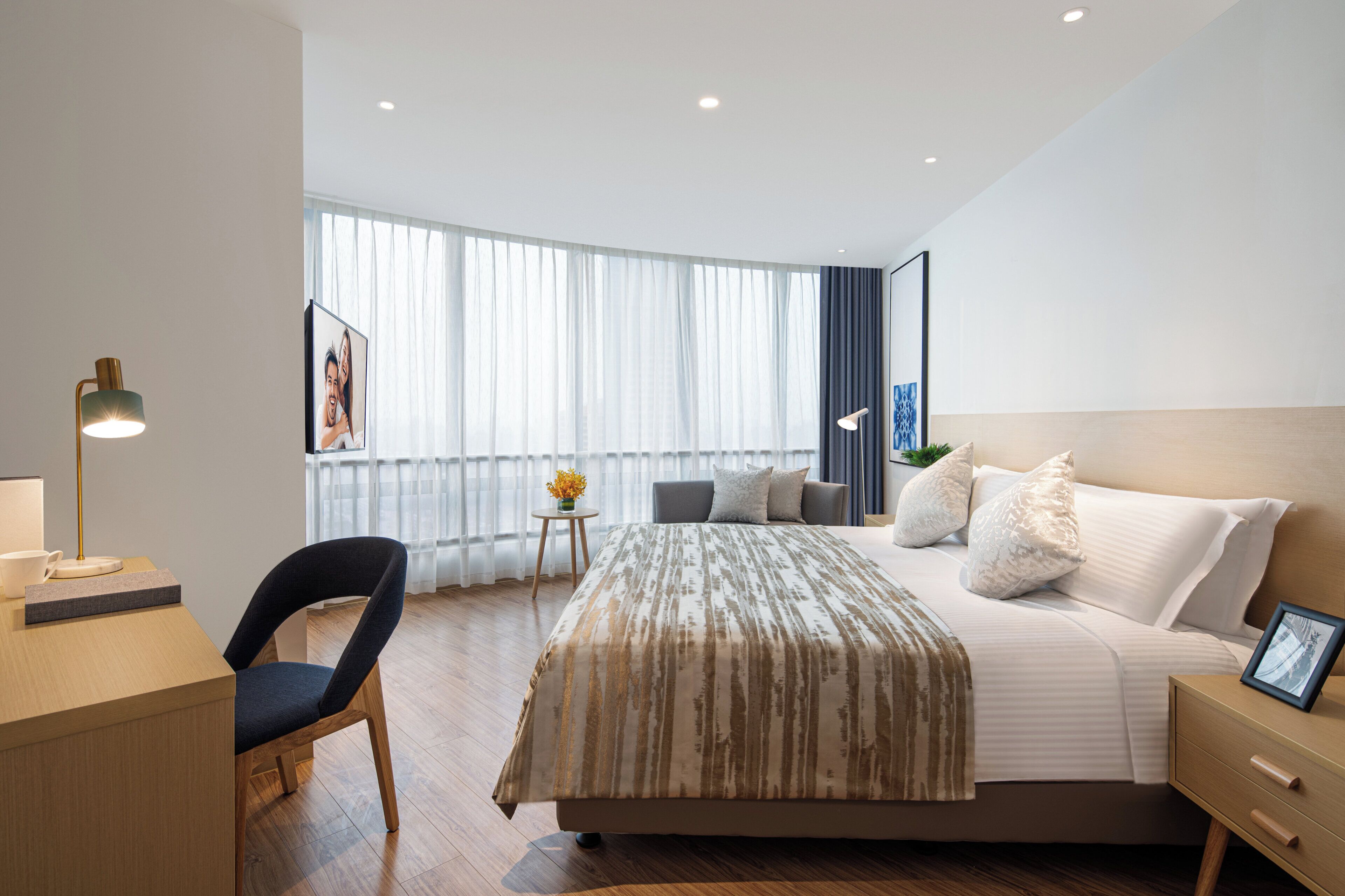 A modern hotel room with a bed, bedside table, chair, and a desk with a lamp.