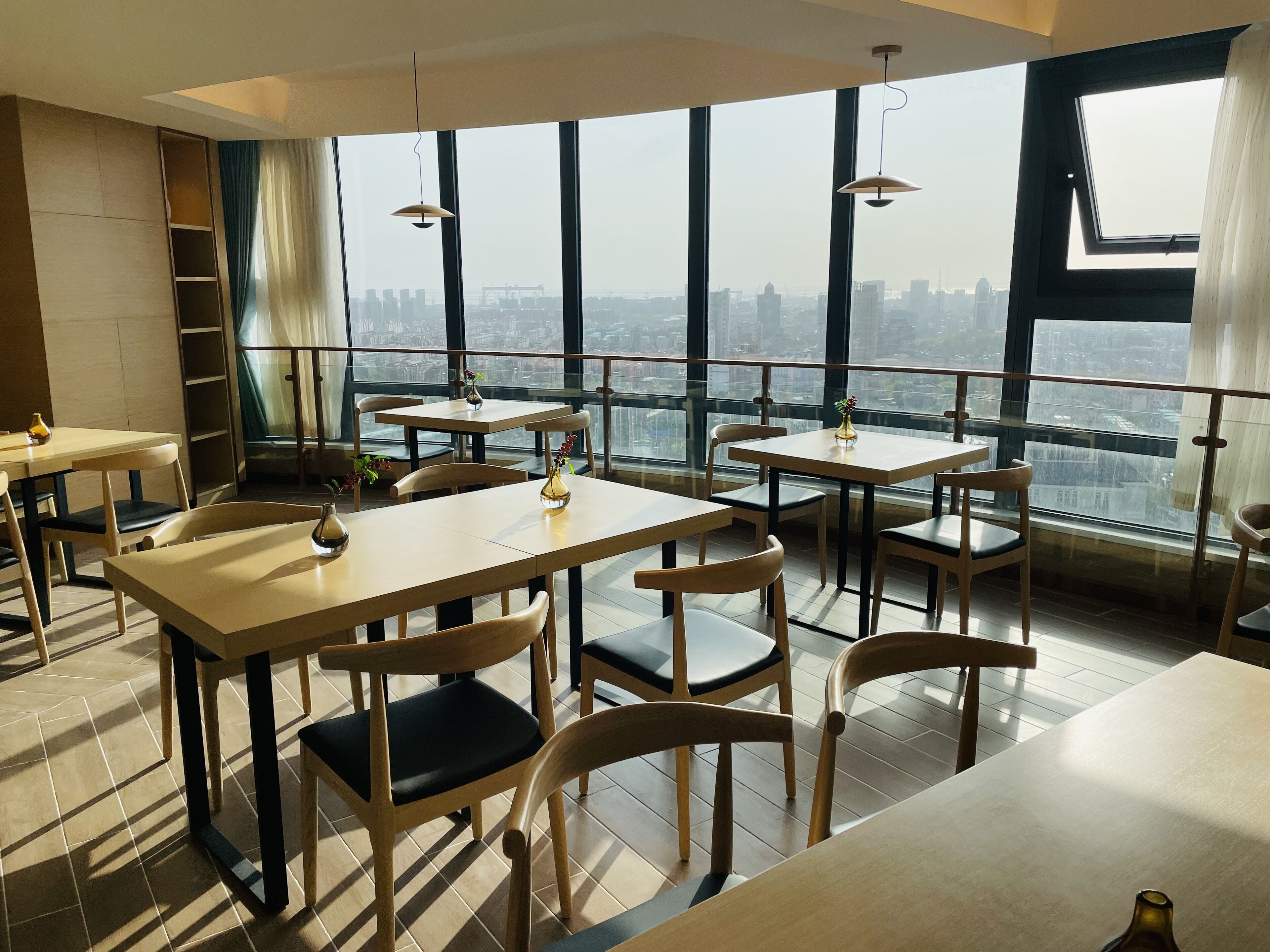 A modern restaurant with high tables and chairs, large windows offering a city view, and pendant lighting.
