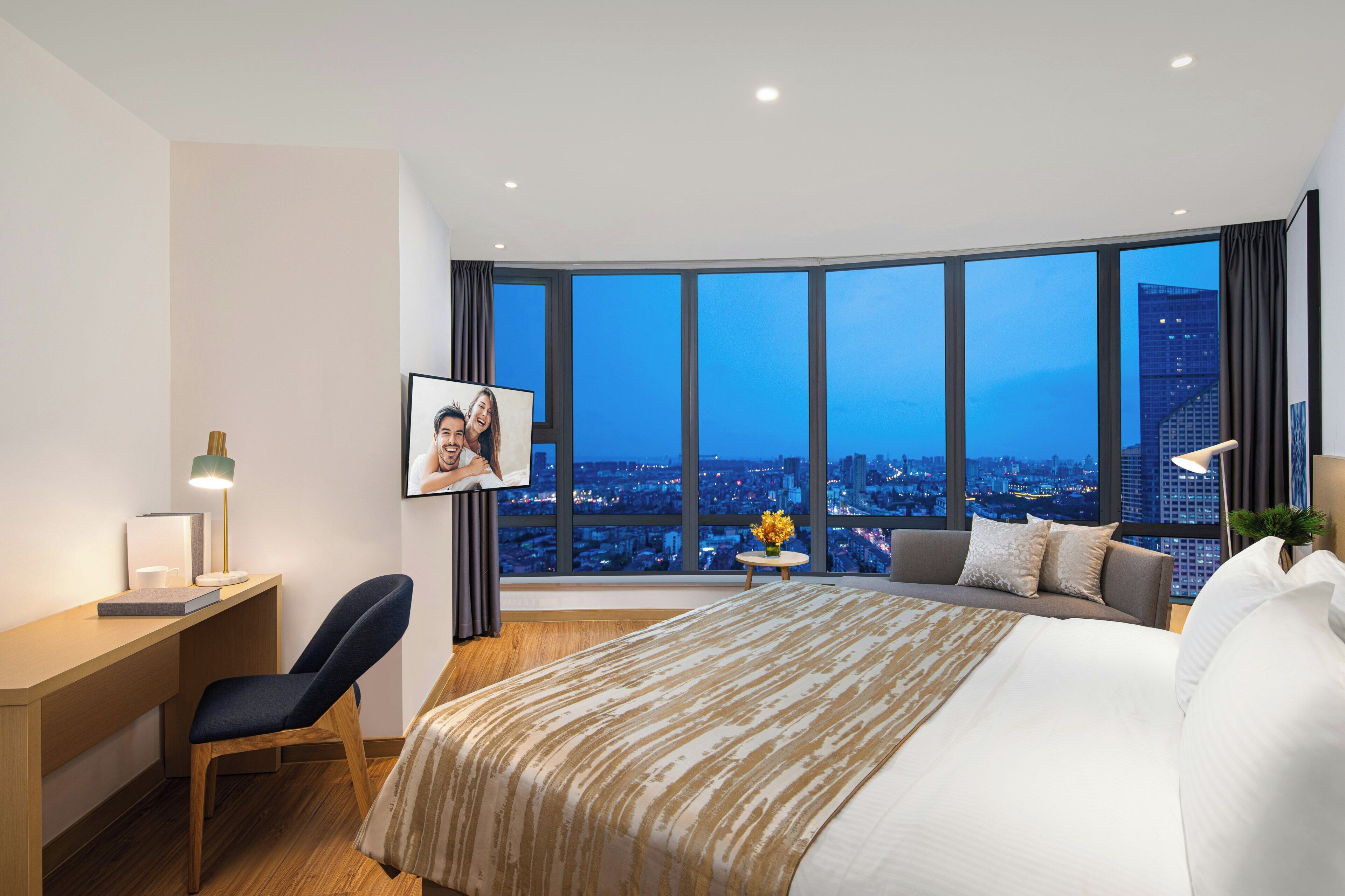 Habitación de hotel con una cama grande, un escritorio con silla, un cuadro en la pared y vistas nocturnas de la ciudad.