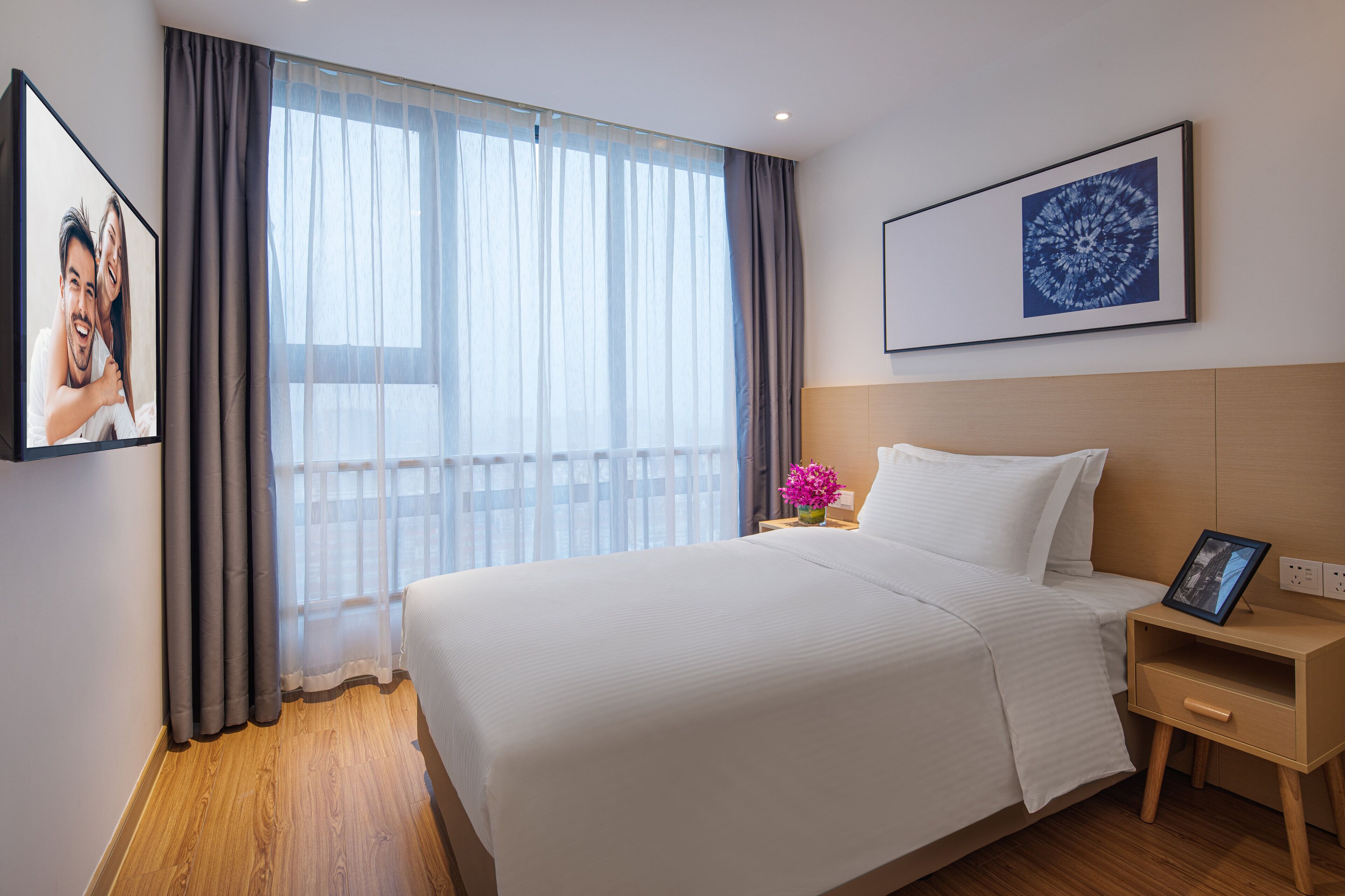 A hotel room with a large bed, a bedside table with a lamp, a picture frame, and a window with sheer curtains.
