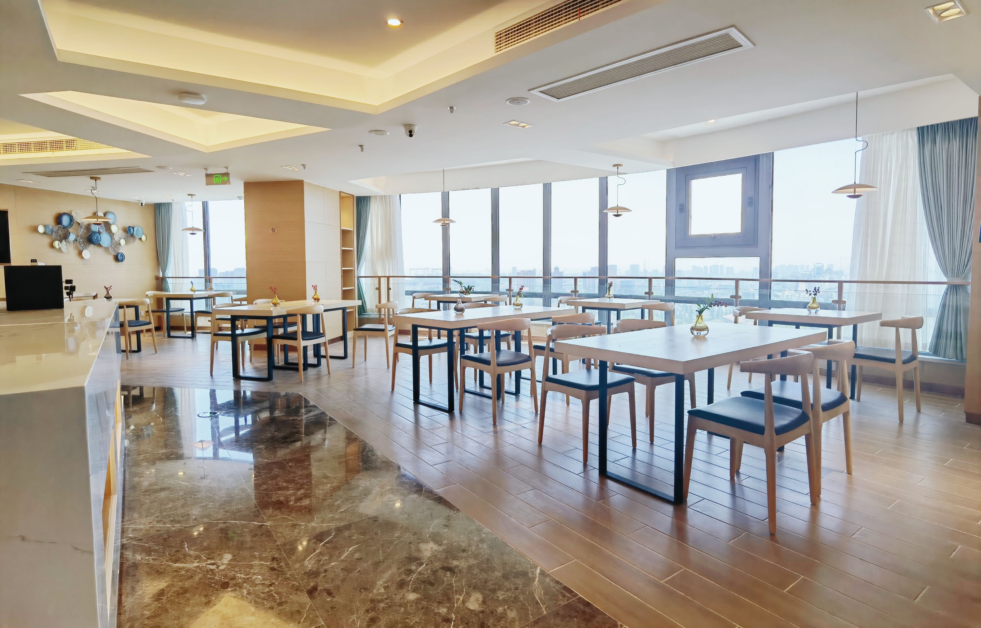 A spacious hotel dining area with multiple tables and chairs, large windows offering city views, and a modern ceiling design.