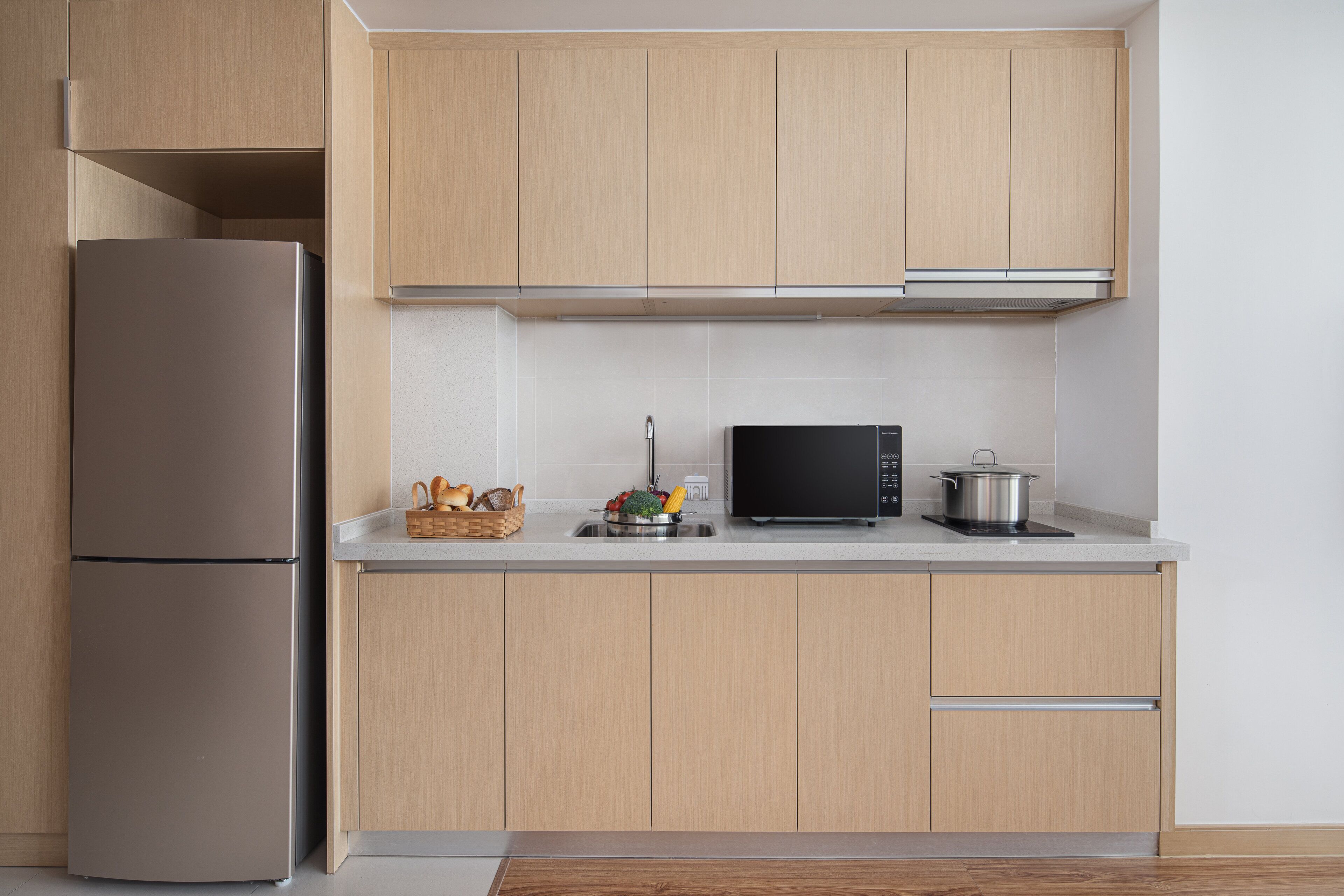 Una cocina moderna con armarios de madera clara, un refrigerador de acero inoxidable, un microondas y una zona de fregadero con una cesta de pan y verduras.