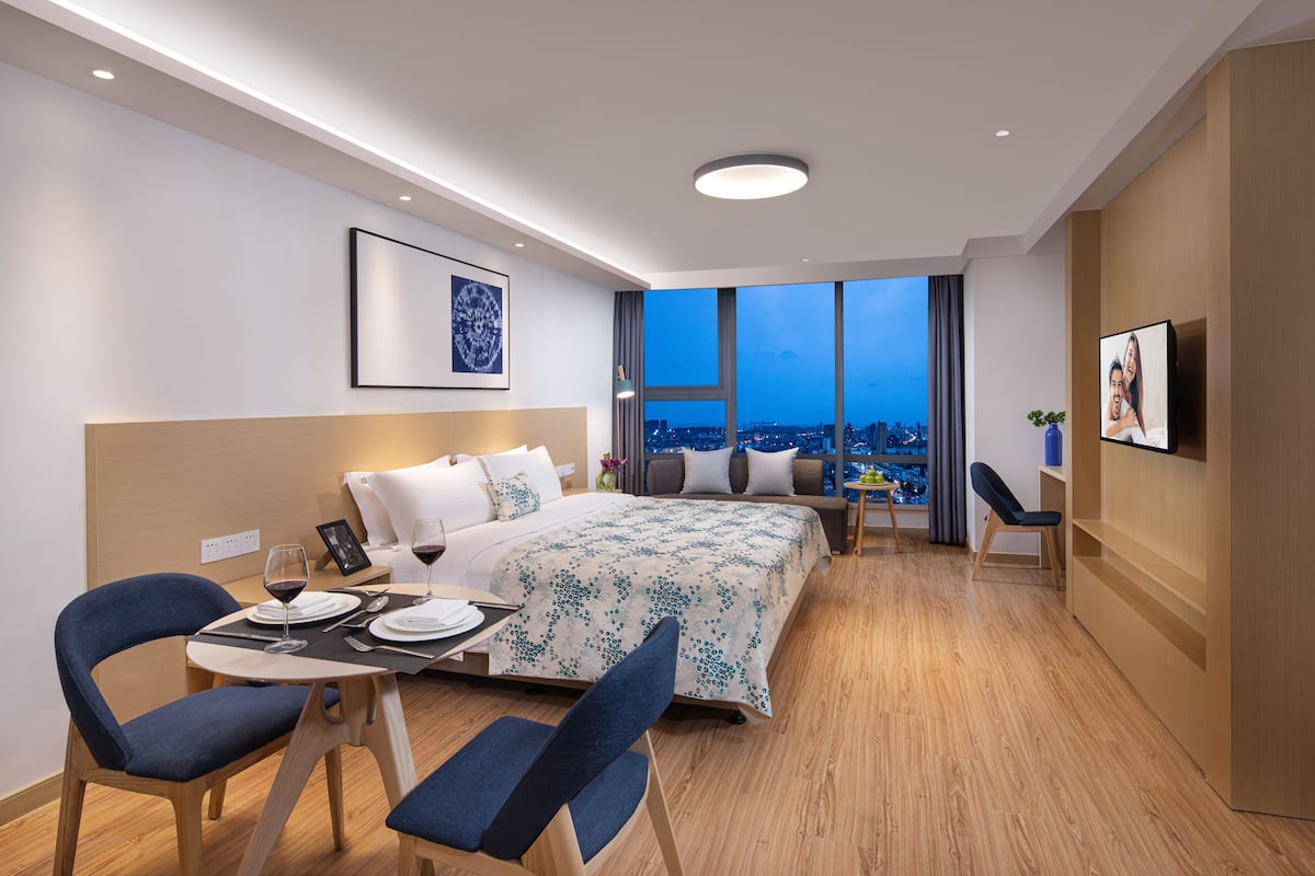 Une chambre d’hôtel moderne dotée d’un grand lit, d’une table à manger avec des chaises, d’un téléviseur et offrant une vue sur la ville grâce à de grandes fenêtres.