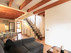 A modern living room with a grey sofa, a dining area with a table and chairs, a kitchen with a refrigerator, and a wooden staircase. - Eu Forat: Townhouse centrally located in Pollensa (Pollença)