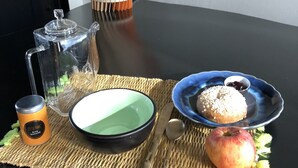 A table with a clear glass pitcher, a black bowl, a blue plate with a pastry, a small cup, a spoon, and an apple on a woven placemat. - Villa Saint-Armand - Chambres d'hotes (Salles)