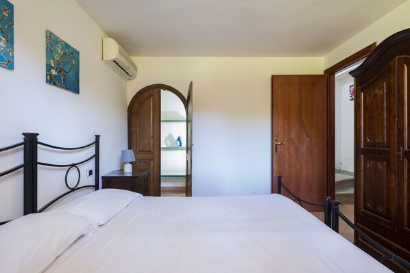 A bedroom with a bed, a wooden wardrobe, a nightstand with a lamp, and a door leading to another room. - Olivastro Holiday Home (Tortoli')