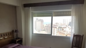 A hotel room with a large bed, a wooden nightstand with a lamp, a chair, a small table, and a window with a city view. - Apartamento Las Torres Con Vista Al Mar (Fuengirola)