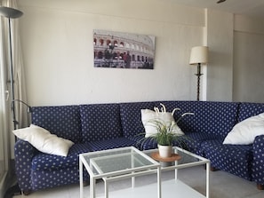 Een woonkamer met een blauwe bank, een glazen salontafel, een lamp en een foto van het Colosseum aan de muur.