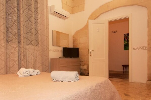 A hotel room with a bed, a TV on a wooden dresser, a door leading to another room, and patterned curtains. - Wish Rooms (Lecce)