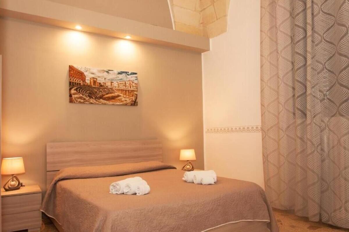 A hotel room with a bed, two bedside lamps, a painting on the wall, and a patterned curtain.