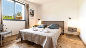 A neatly arranged bedroom with a bed, a chair, a nightstand, and a window with a view. - Charming apartments in Porto-Vecchio and Araggio, between the sea and the mounta (PORTO VECCHIO)