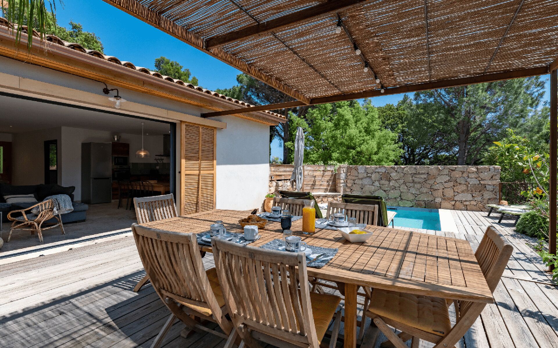 Un patio al aire libre con mesa y sillas de madera, un muro de piedra y una piscina.