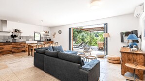 A modern living room with a grey sofa, wooden dining table, and chairs. A kitchen with a black countertop and wooden cabinets is visible. There is a TV mounted on the wall and a window providing a view of the outdoors. - Charming apartments in Porto-Vecchio and Araggio, between the sea and the mounta (PORTO VECCHIO)