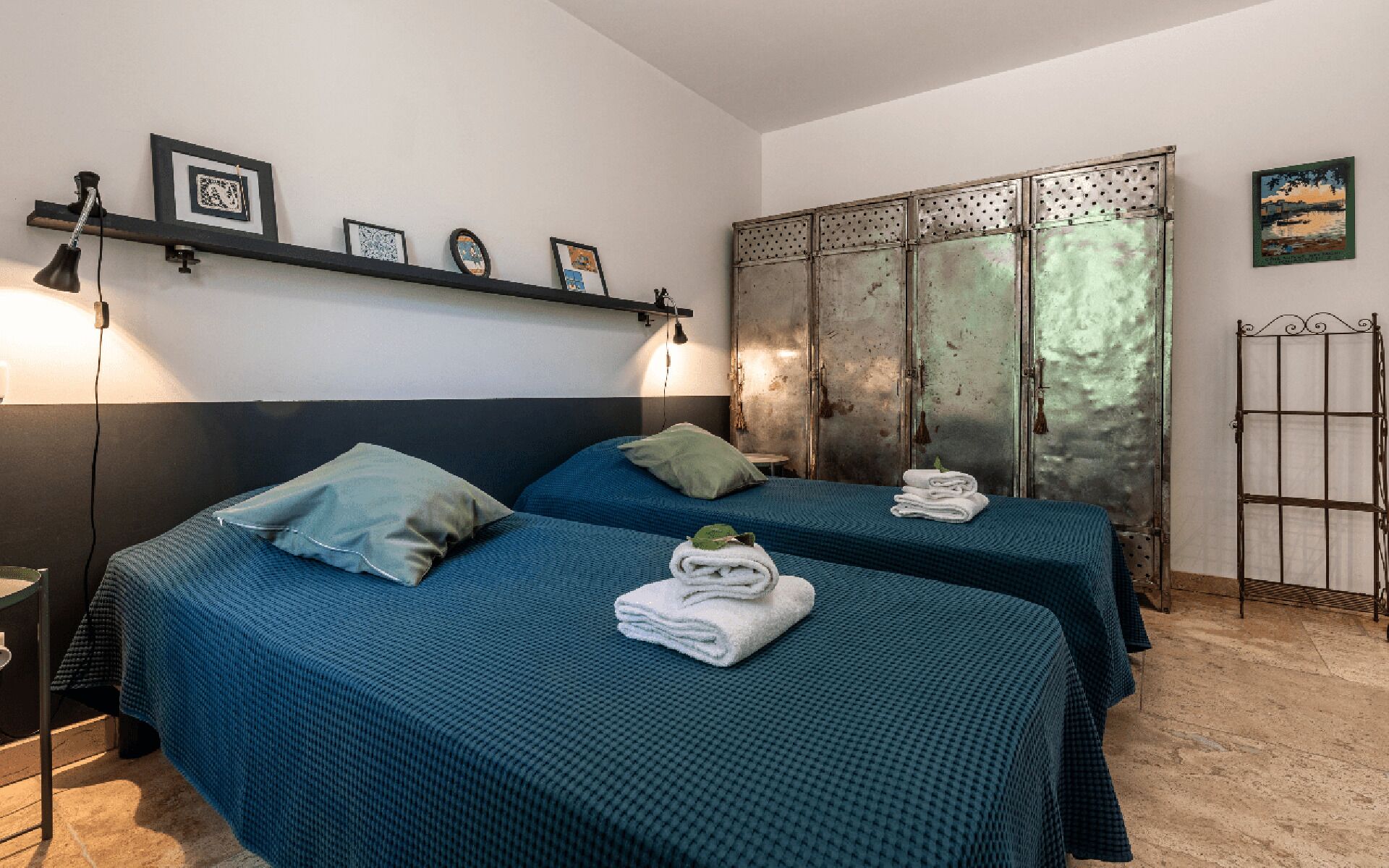 A room with two beds, each with a blue bedspread and pillows, a metal wardrobe, and a shelf with framed pictures.