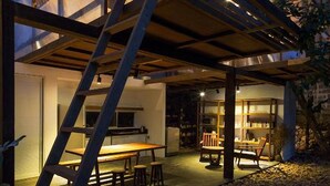Uma casa moderna com fachada de vidro e exterior de madeira. Possui uma escadaria que leva a um andar superior, e uma área de jantar com mesa e cadeiras.