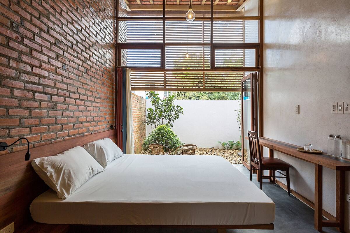 Un dormitorio con pared de ladrillo, una cama con sábanas blancas, un escritorio de madera y vista a un jardín a través de una puerta de vidrio.