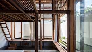 Quarto com teto inclinado, vigas de madeira, cama, escrivaninha e janelas amplas.