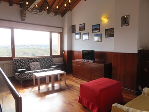 Ein Wohnzimmer mit einer Couch, einem Tisch, einem Fernseher auf einer Holzkommode und einem roten Hocker. An der Wand befinden sich gerahmte Bilder und ein großes Fenster mit Aussicht.