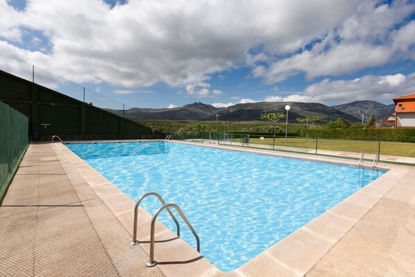 Ein Swimmingpool mit klarem, blauem Wasser, umgeben von einer Betonplattform und einem grünen Zaun. Im Hintergrund erheben sich Berge und der Himmel ist wolkenlos.