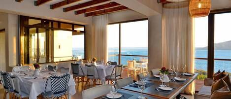 Un espace repas avec plusieurs tables dressées pour le repas, de grandes fenêtres offrant une vue sur la mer et un plafond haut avec des poutres en bois apparentes.