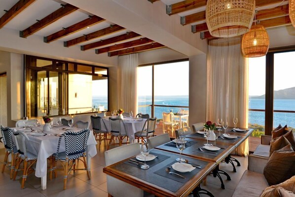 Un comedor con varias mesas dispuestas para una comida, ventanas amplias con vistas al mar y un techo alto con vigas de madera a la vista.