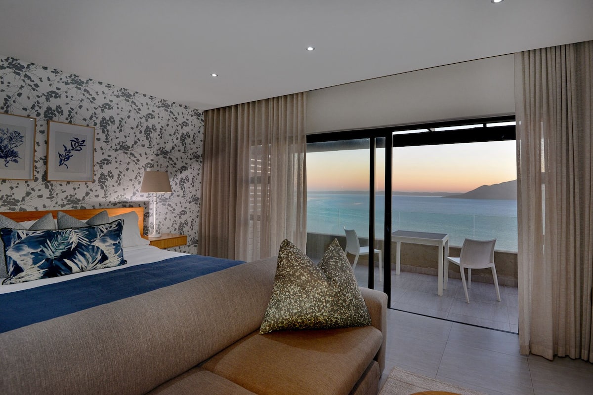 A hotel room with a bed, a sofa, a balcony with a view of the sea, and a dining area.