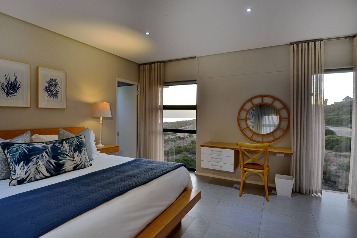 A bedroom with a bed, a desk, a chair, a window with curtains, and a view of the outdoors.
