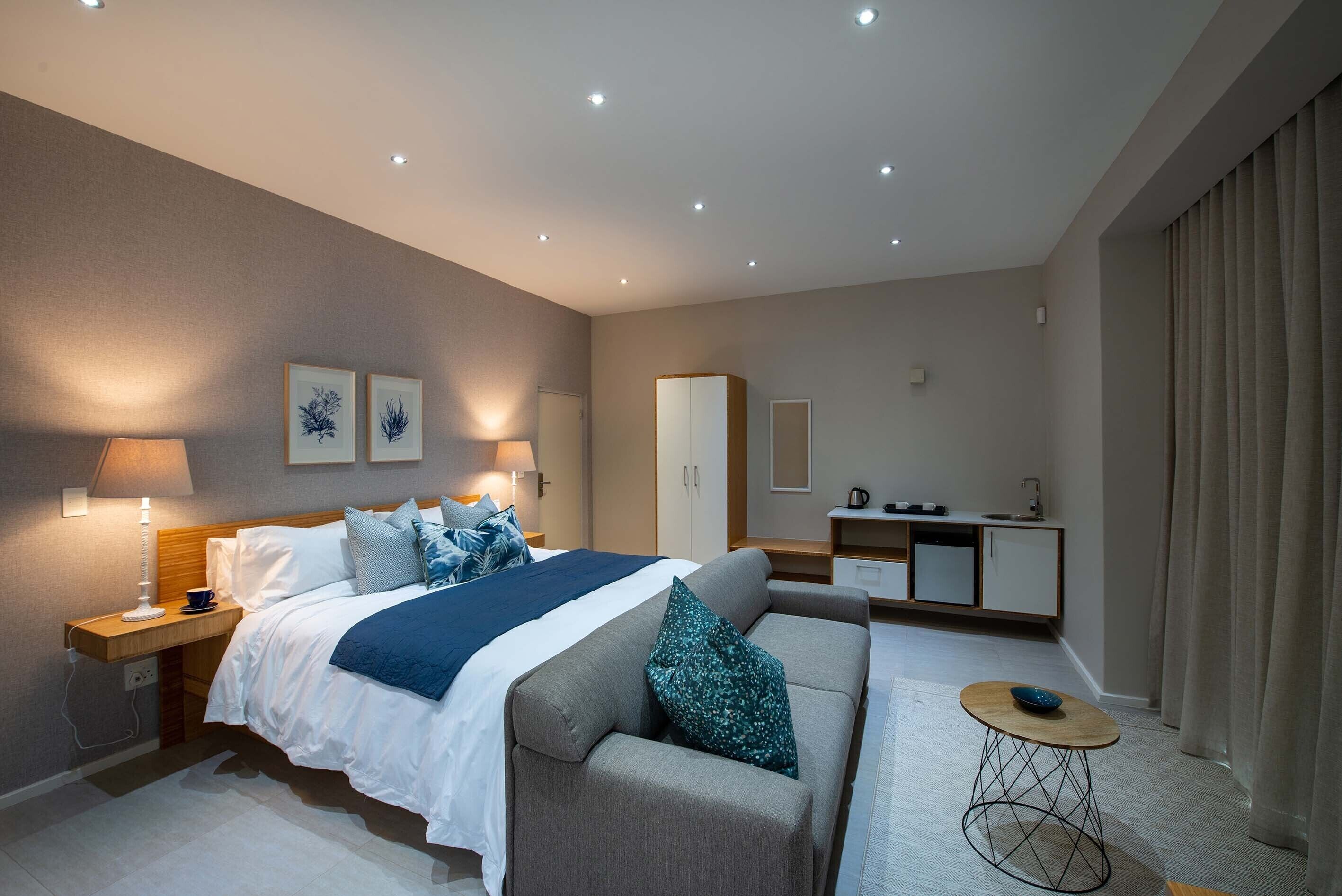 Una habitación de hotel moderna con cama, sofá, mesita de noche, lámpara y una pequeña zona de kitchenette.