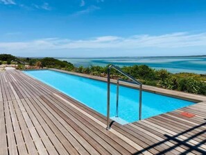 Una piscina rectangular con muelle de madera, barandales de metal y vistas al océano.