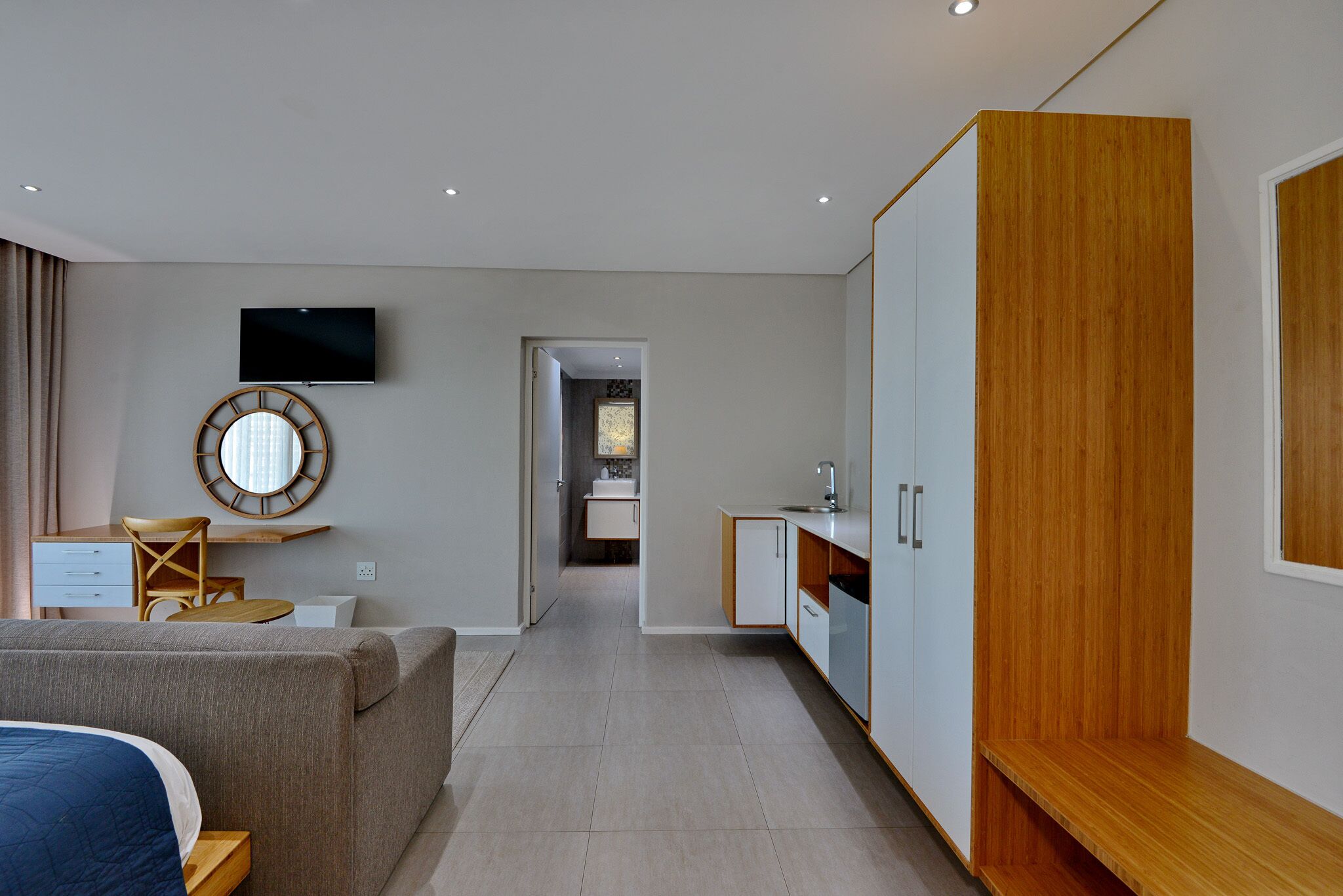 A modern hotel room with a bed, a desk, a TV, a mirror, and a kitchenette.