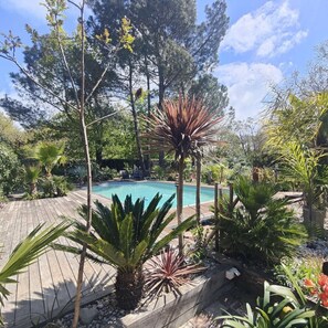A pool surrounded by tropical plants and trees, with a wooden deck and a stone wall. - La Grange Océane Lège Cap-Ferret - "Douceur Marine" Studio - Heated Pool - WIFI/netflix (LEGE CAP FERRET)