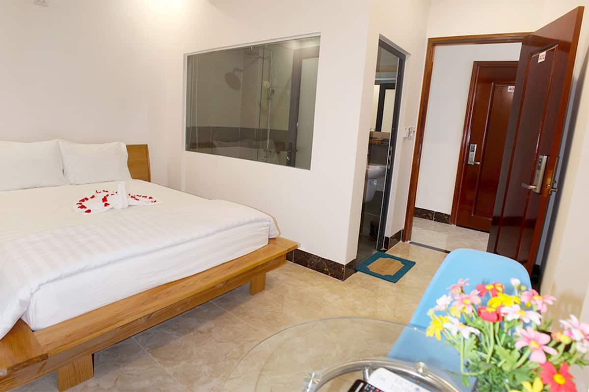 Habitación de hotel con cama de madera, ropa blanca, cabina de ducha de vidrio y una puerta con número.