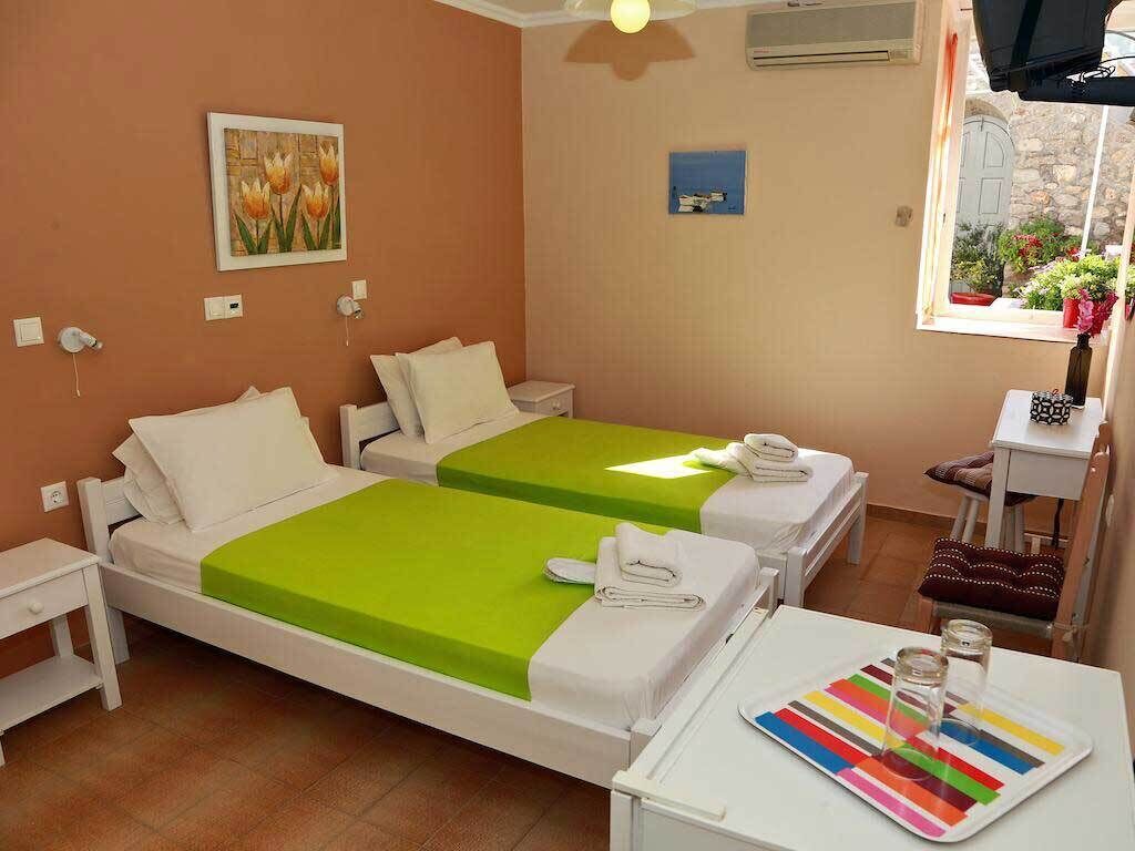 A hotel room with two beds, a small table with a tray of glasses, a window with plants, and a painting on the wall.