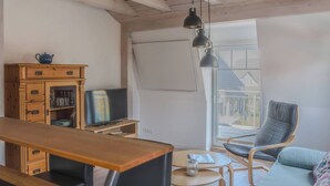 A room with a wooden desk, a television, a sofa, and a chair. There is a window with a view of a house. - Apartments with a large balcony near the beach (Bosau)