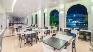 A spacious indoor dining area with multiple tables and chairs, large windows with a view of the street at night, and a ceiling fan. - Vapa Hotel (Phuket)