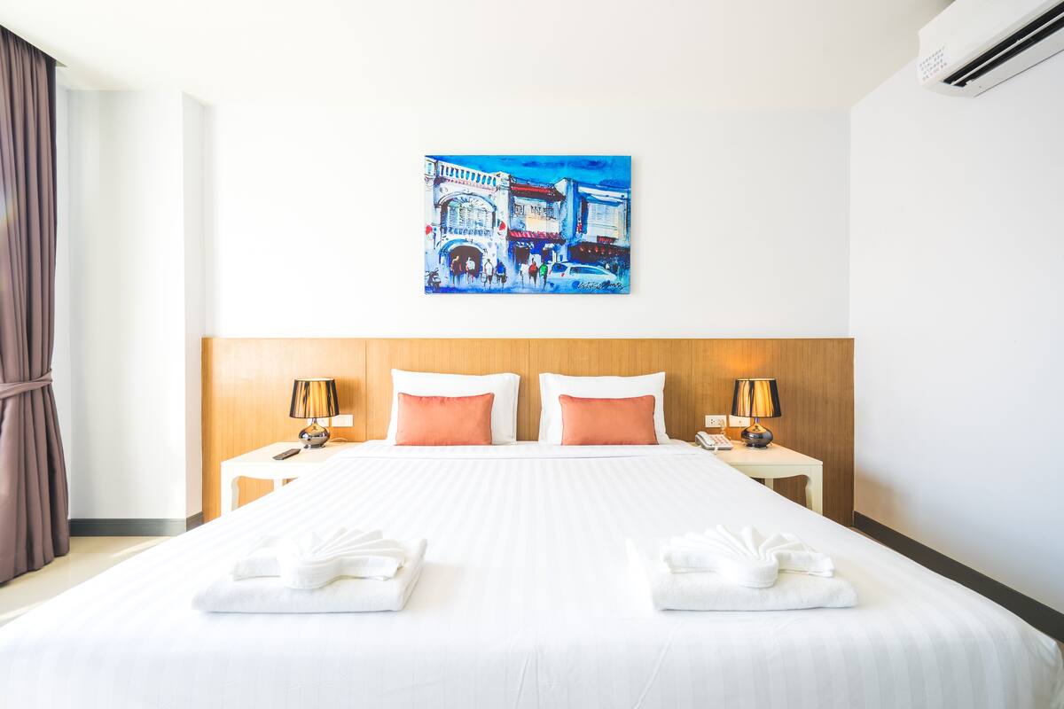 A hotel room with a large bed, two bedside tables with lamps, a painting on the wall, and a view of the street outside.