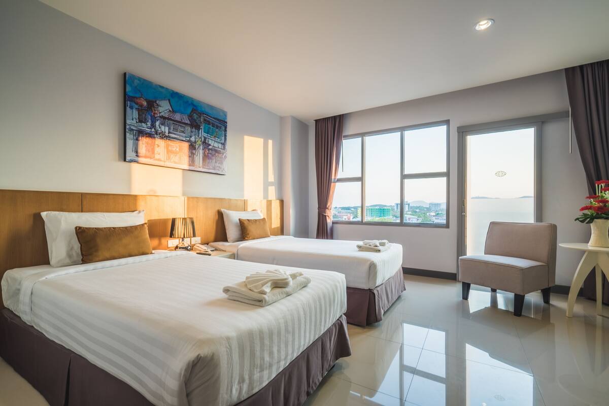 A hotel room with two beds, a desk, a chair, and a large window offering a city view.