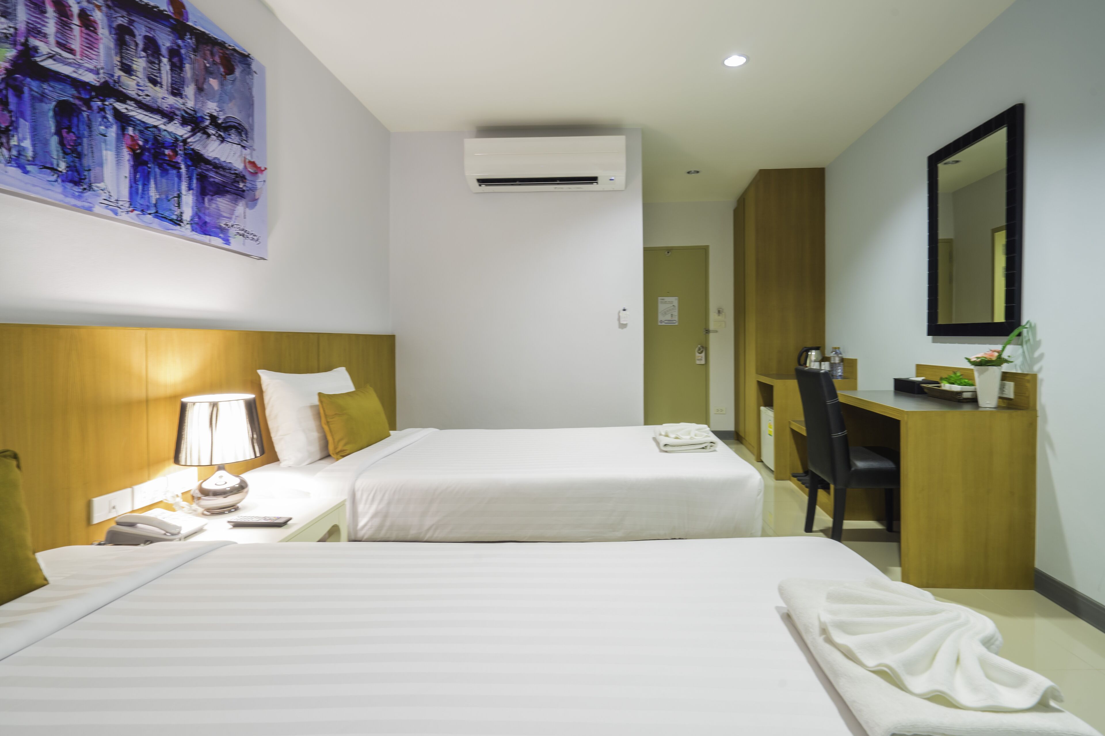 A hotel room with two beds, a desk, a chair, a lamp, an air conditioner, and a painting on the wall.