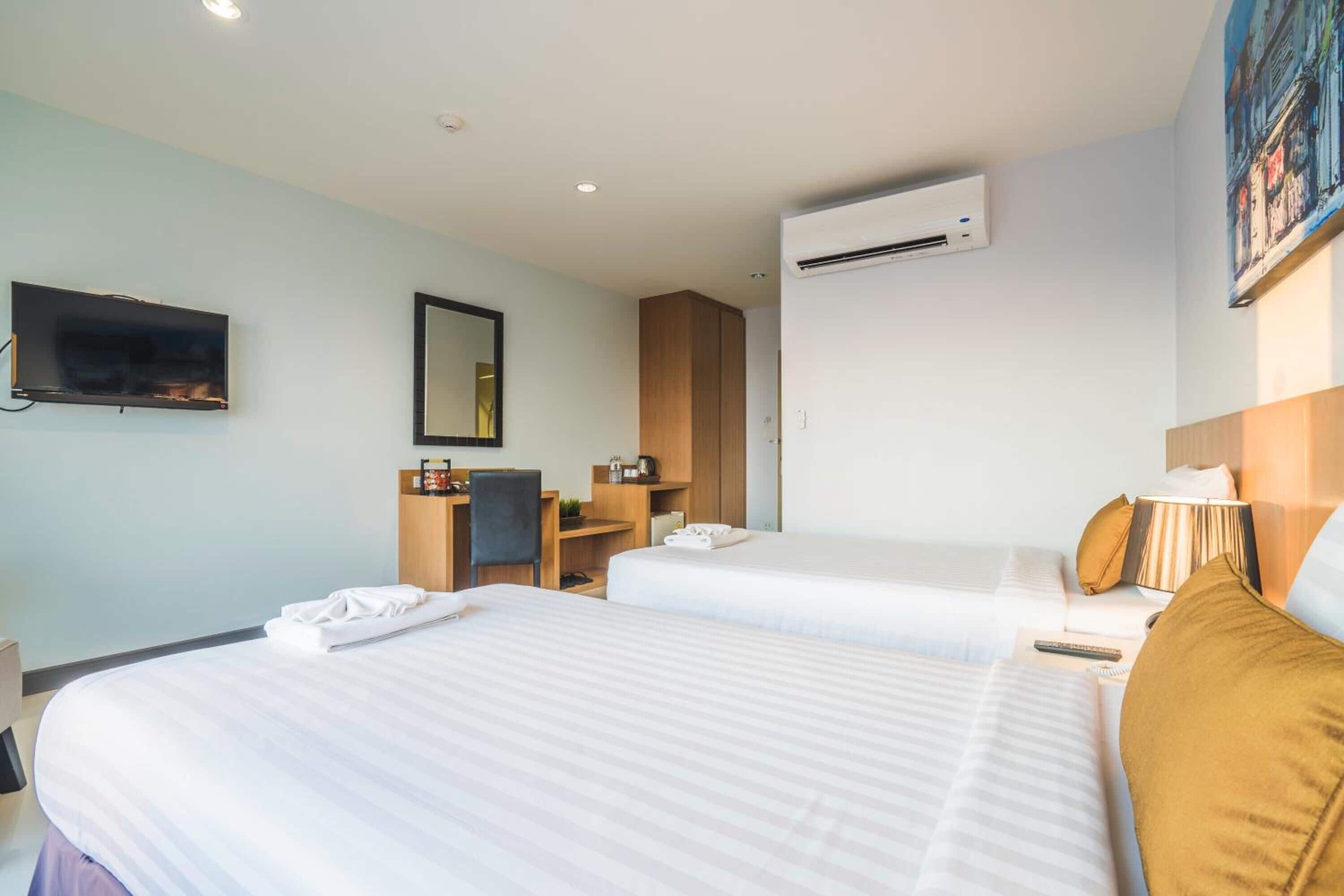 A hotel room with two beds, a desk, a chair, a TV, and an air conditioning unit.